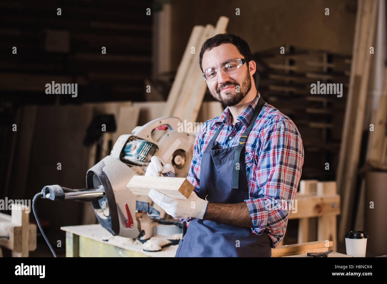 Beau charpentier de lunettes de protection est à la caméra et au sourire alors qu'il se tenait près de son objet en bois dans l'atelier Banque D'Images