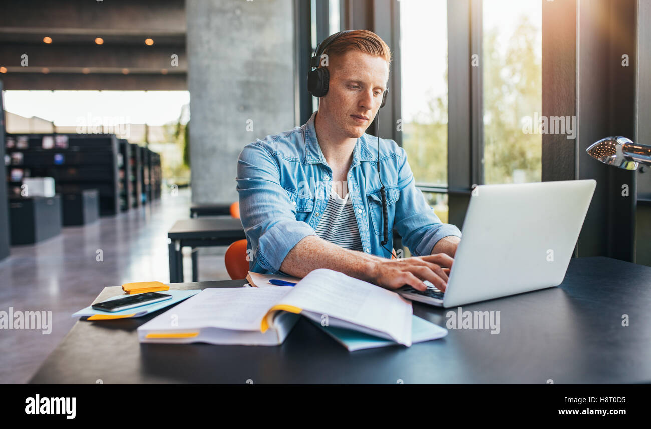 Beau mâle étudiant dans une bibliothèque universitaire travaillant sur ordinateur portable. Jeune homme l'étude sur l'affectation de l'école. Banque D'Images