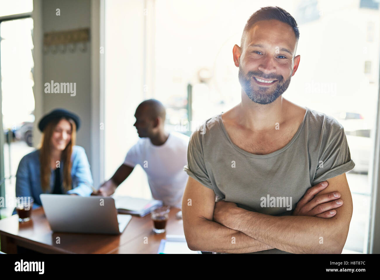 Handsome smiling barbu en chemise à manches courtes, les bras croisés et l'article dans le café avec les clients en arrière-plan Banque D'Images