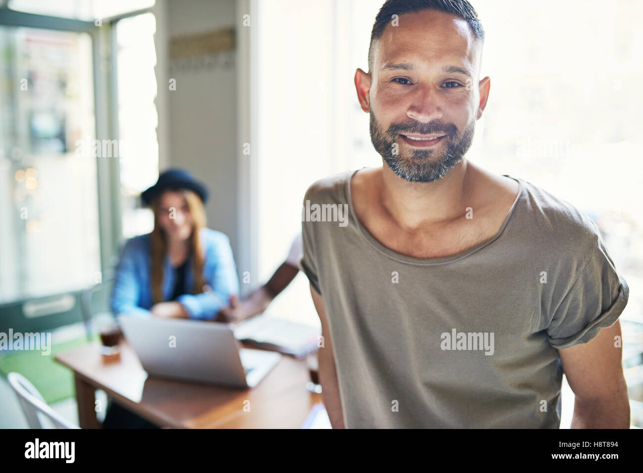 Souriant beau barbu en short sleeve shirt leaning on table in coffee house with woman in background Banque D'Images