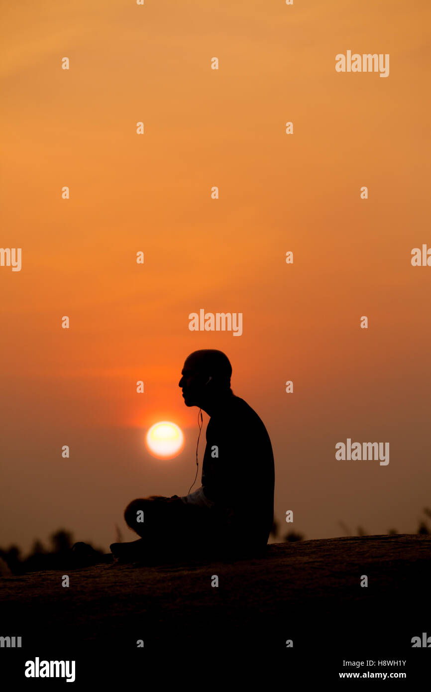 Mâle adulte photographié silhouetté contre un coucher de soleil, avec écouteur et fils montrant, écouter de la musique et la méditation Banque D'Images