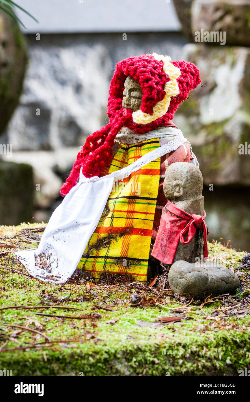 Le Japon, Koyasan, Okunoin cemetery. Les Jizo bosatsu assis statue bouddhiste avec soumission jaune et rouge Chapeau tricoté assis sur la mousse en face de tombstone. Banque D'Images
