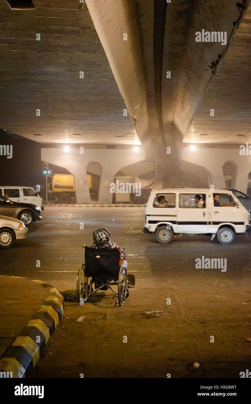 Un vieil homme dans un fauteuil roulant à la mendicité par une route très fréquentée à Nizamuddin à New Delhi. Banque D'Images