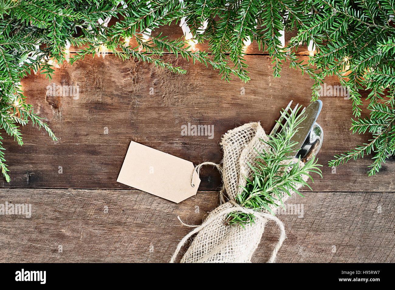 Arbre de Noël des branches de pins, de l'argenterie enveloppée dans une serviette de toile avec carte vierge et des lumières décoratives sur un fond rustique en bois de grange. Banque D'Images