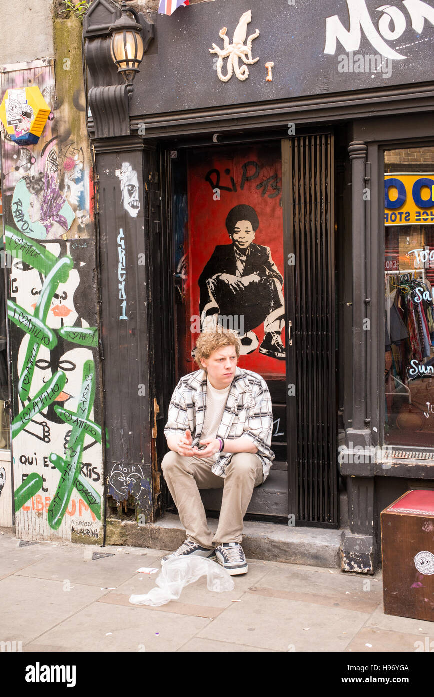 Jeune homme assis sur la rue en face d'un atelier fermé avec mur recouvert de graffitis et des gribouillis dans Brick Lane, Shoreditch, London, UK. Banque D'Images