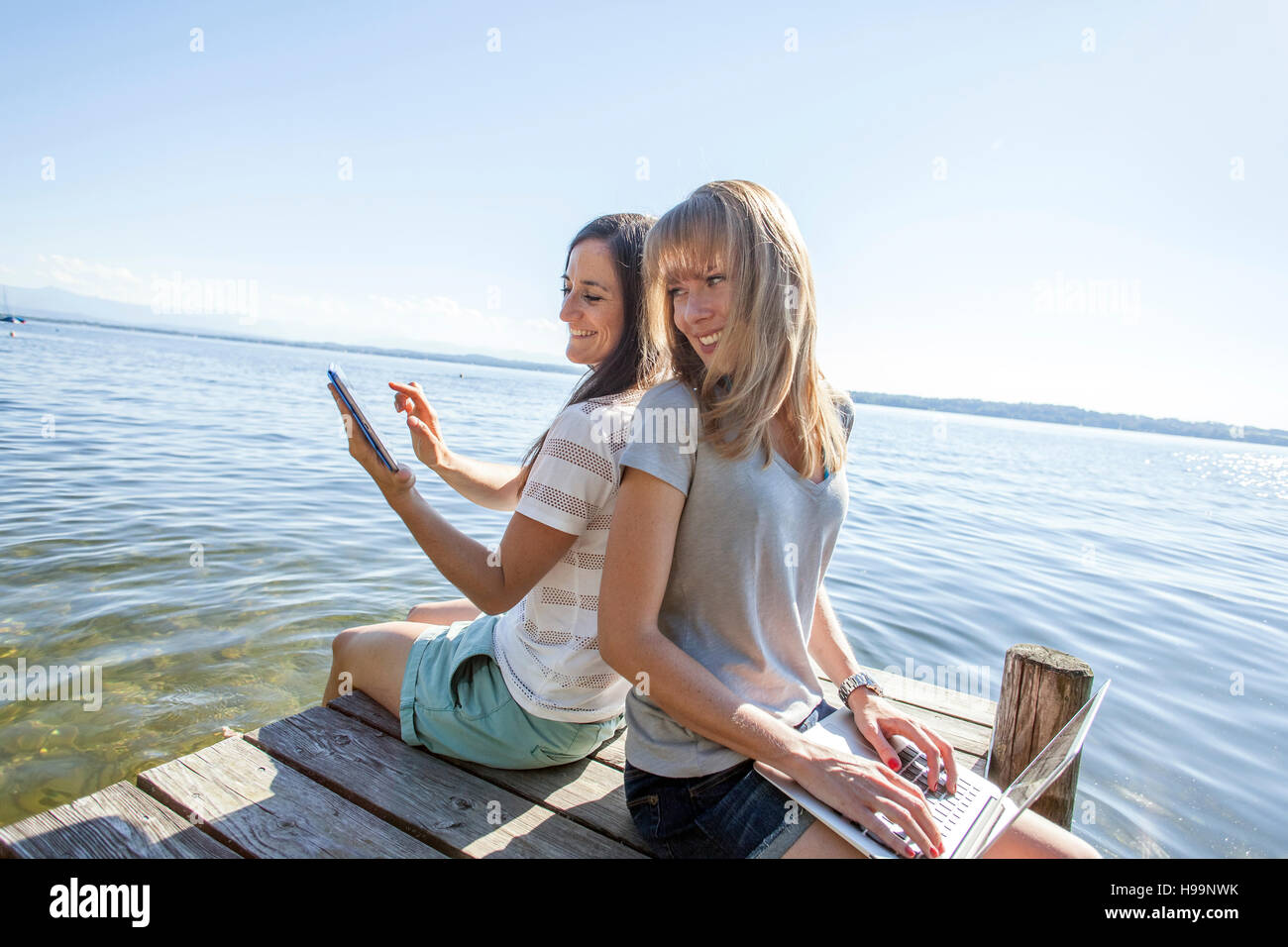 Deux jeunes femmes sont assises sur la jetée par le lac à l'aide d'ordinateur portable et tablette Banque D'Images