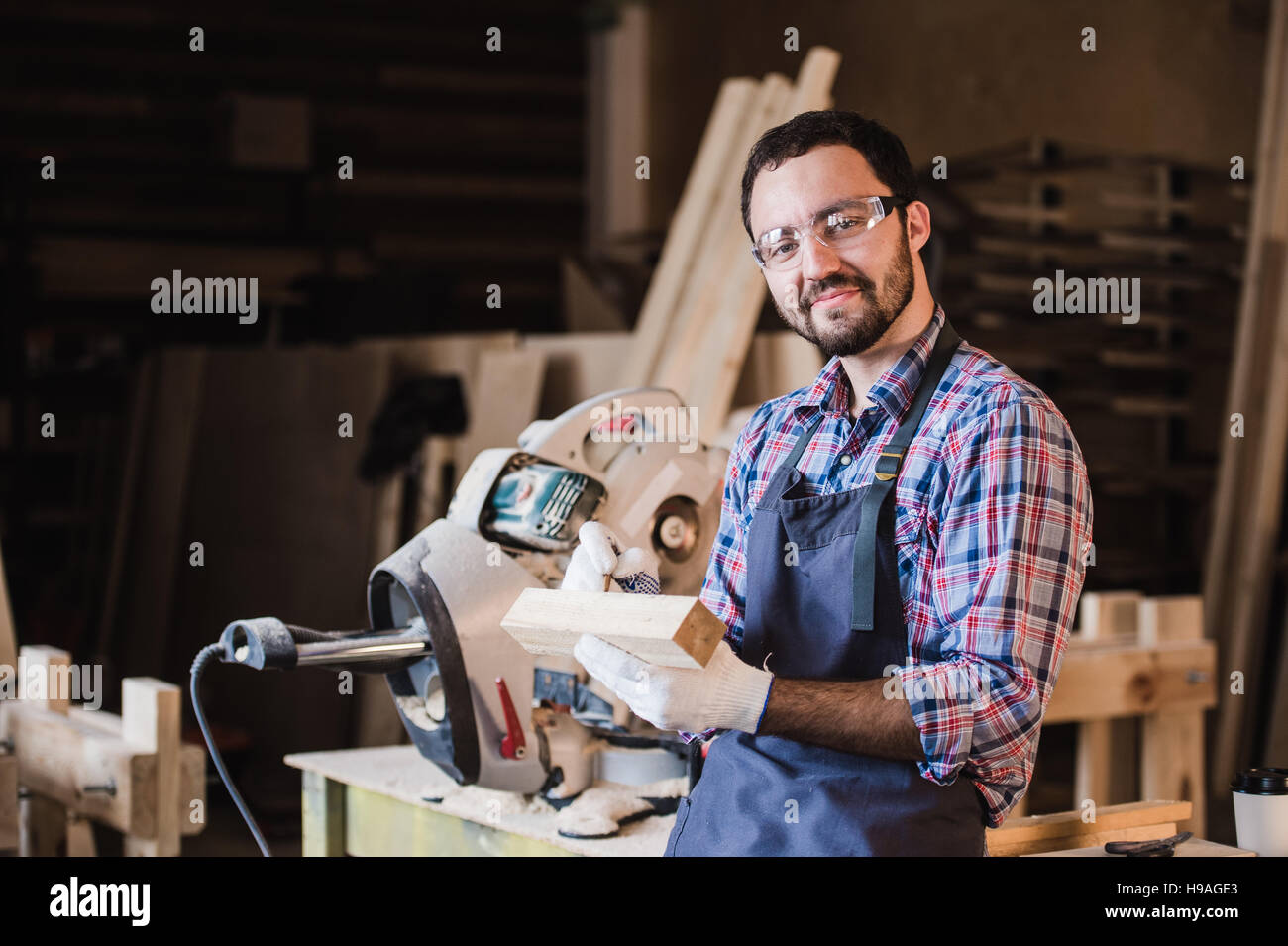 Beau charpentier de lunettes de protection est à la caméra et au sourire alors qu'il se tenait près de son objet en bois dans l'atelier Banque D'Images