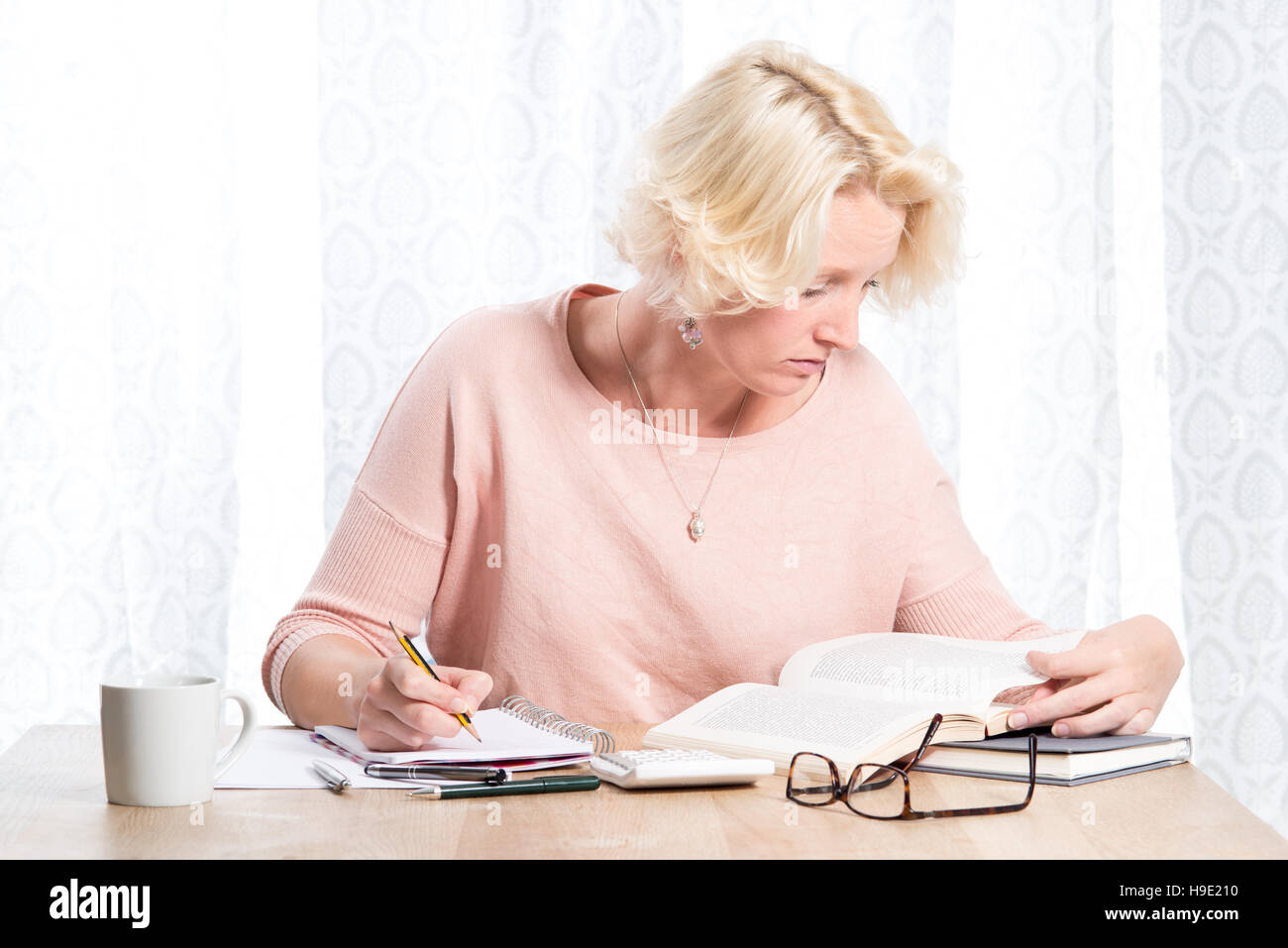 Une femme blonde vêtue d'une légère cavalier rose avec collier étudie un livre tout en écrivant dans un tampon avec un crayon. Elle s'assoit à une table en bois en Banque D'Images