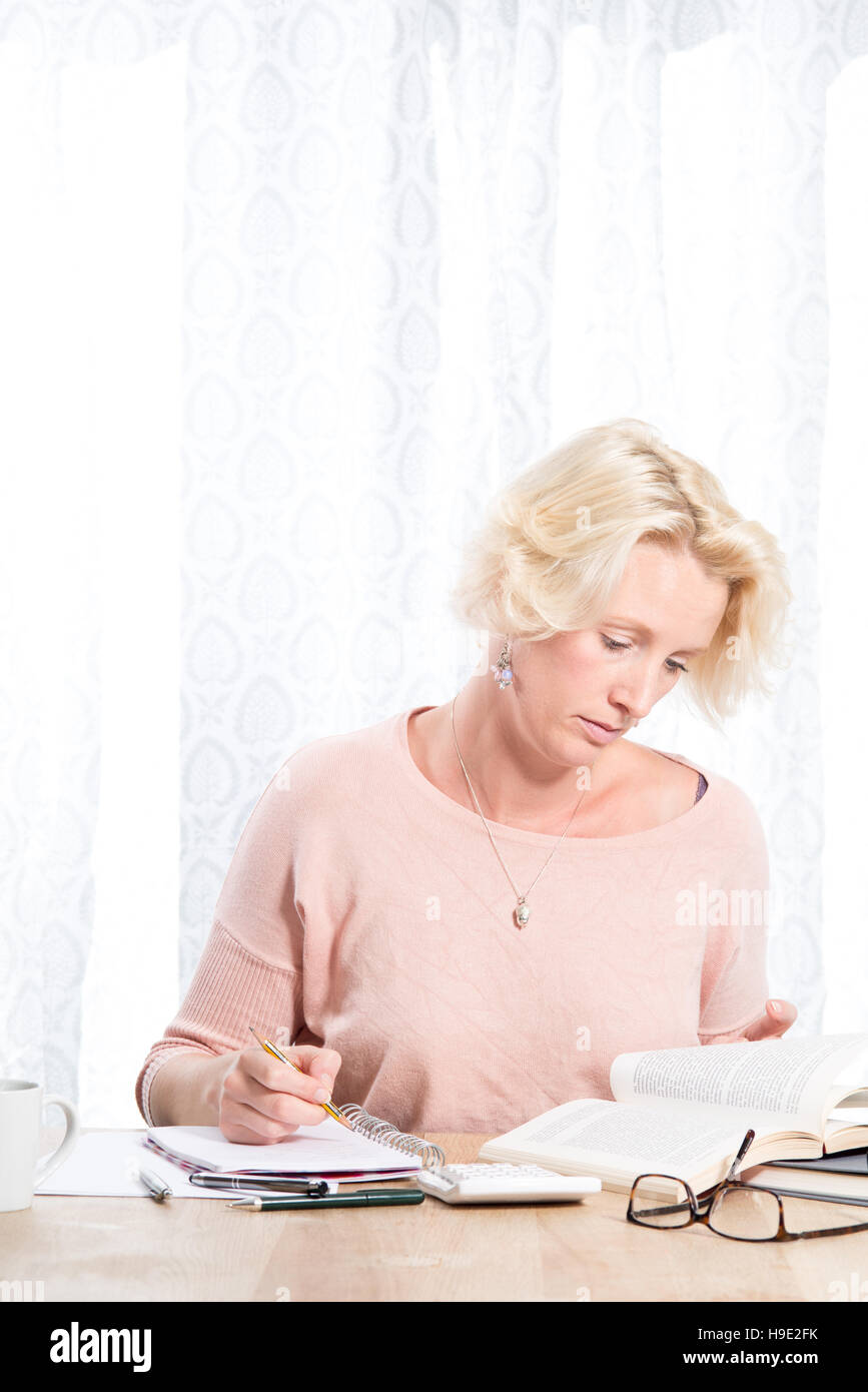 Portrait d'une femme blonde portant un cardigan rose assis à la maison travailler à un bureau en bois. Elle est titulaire d'un crayon pour un pavé d'écriture tout en lisant f Banque D'Images