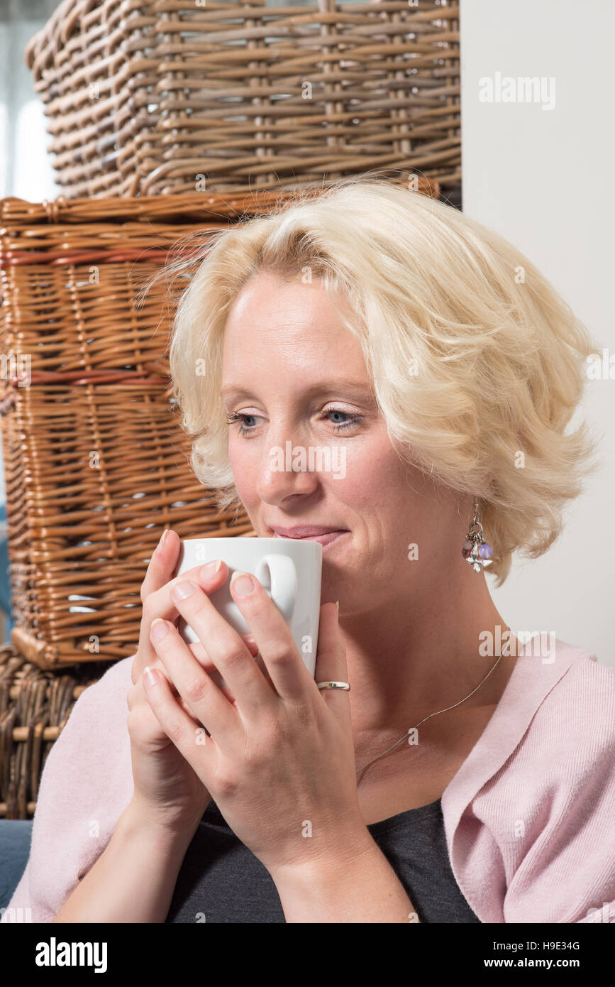 Une femme blonde d'âge moyen se trouve à côté des boîtes en osier sur un canapé ou un canapé, tenant une tasse à deux mains près de sa bouche. Elle sourit avec satisfaction Banque D'Images