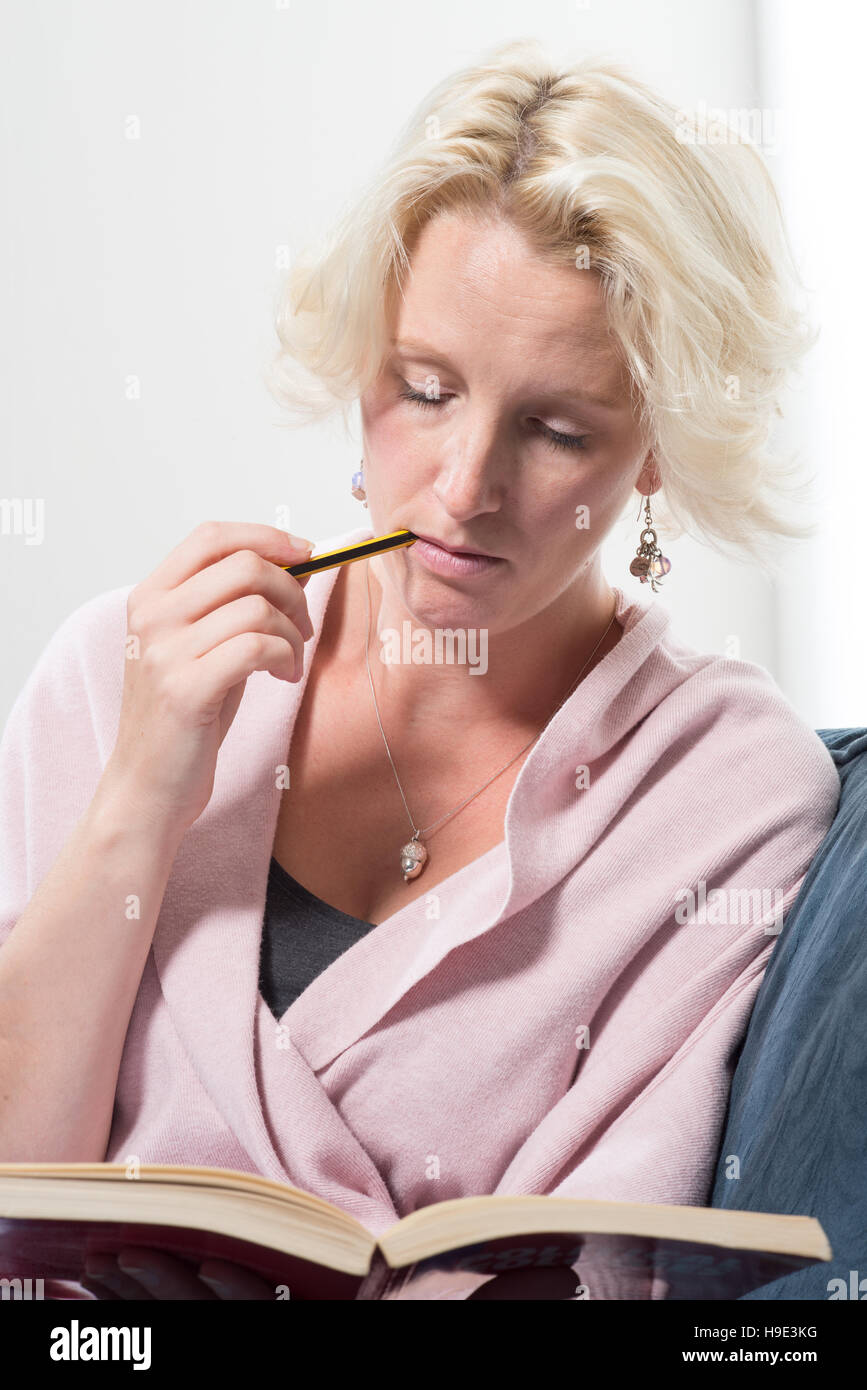 Une femme blonde d'âge moyen habillé est assis sur un canapé ou un canapé bleu tenant un livre et de mordre l'extrémité d'un crayon dans la concentration qu'elle la lit. Banque D'Images