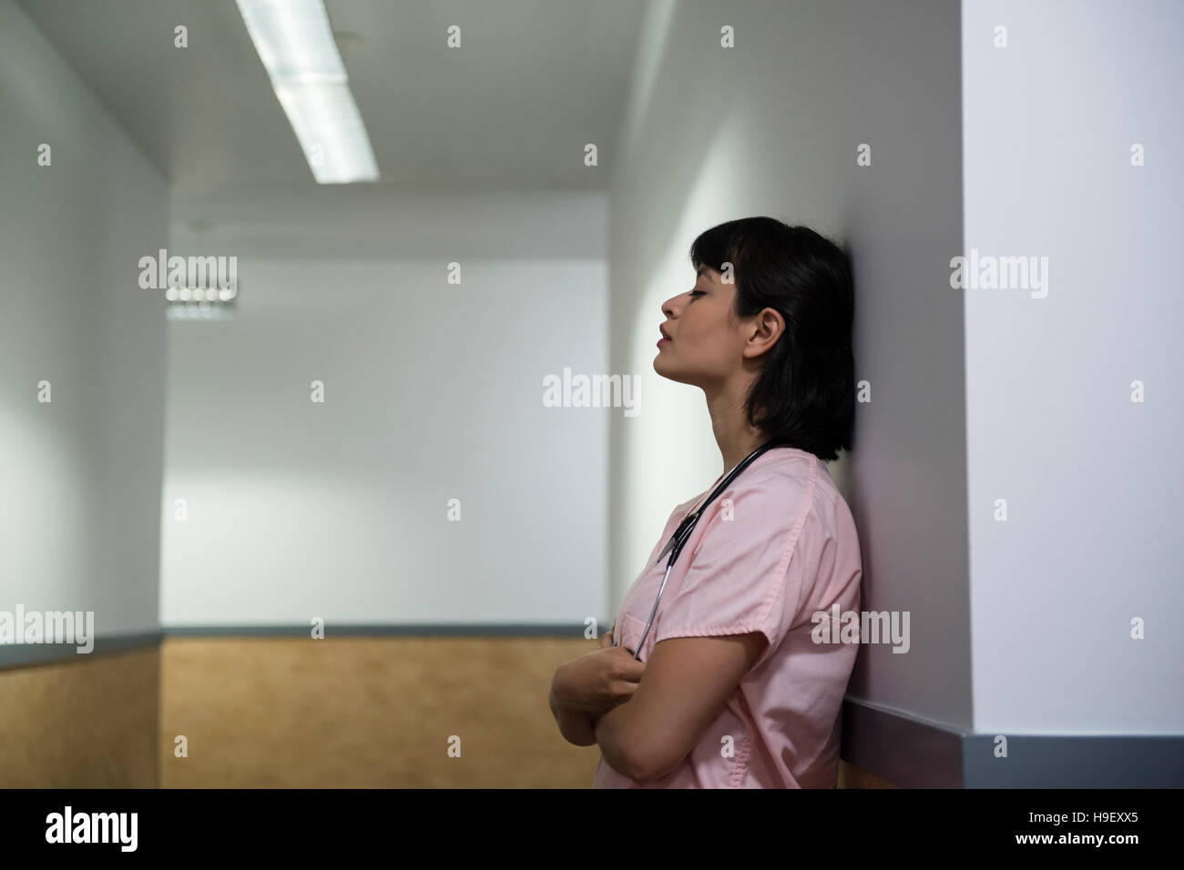 Fatigué Hispanic nurse leaning on wall in hospital corridor Banque D'Images