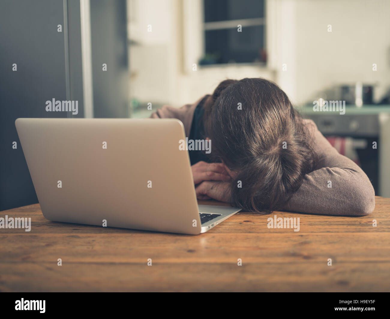 Une jeune femme triste est en train de dormir par son ordinateur portable dans sa cuisine à la maison Banque D'Images