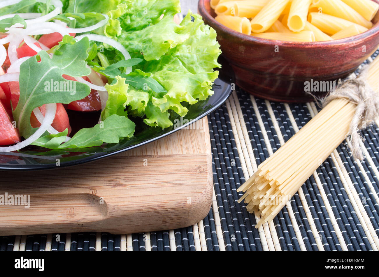 Composition sur la table avec une salade de tomates, oignons, laitue et roquette sur un support en bois, des pâtes et des spaghettis pour la cuisson Banque D'Images