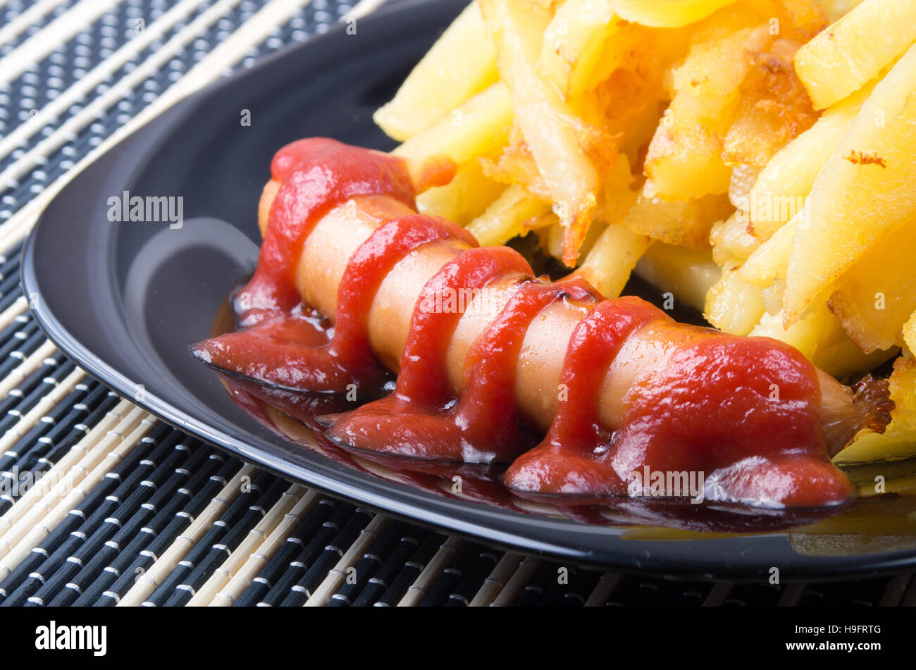 Détail d'une plaque noire avec les saucisses grillées avec sauce tomate et frites close up sur une table à rayures Banque D'Images