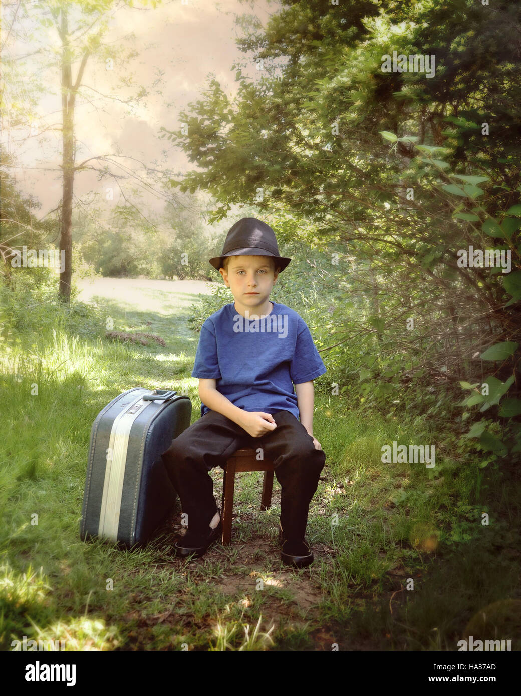 Un jeune garçon est assis sur un sentier dans les bois avec une valise et un chapeau. L'enfant l'air triste pour un voyage ou vacances. Banque D'Images