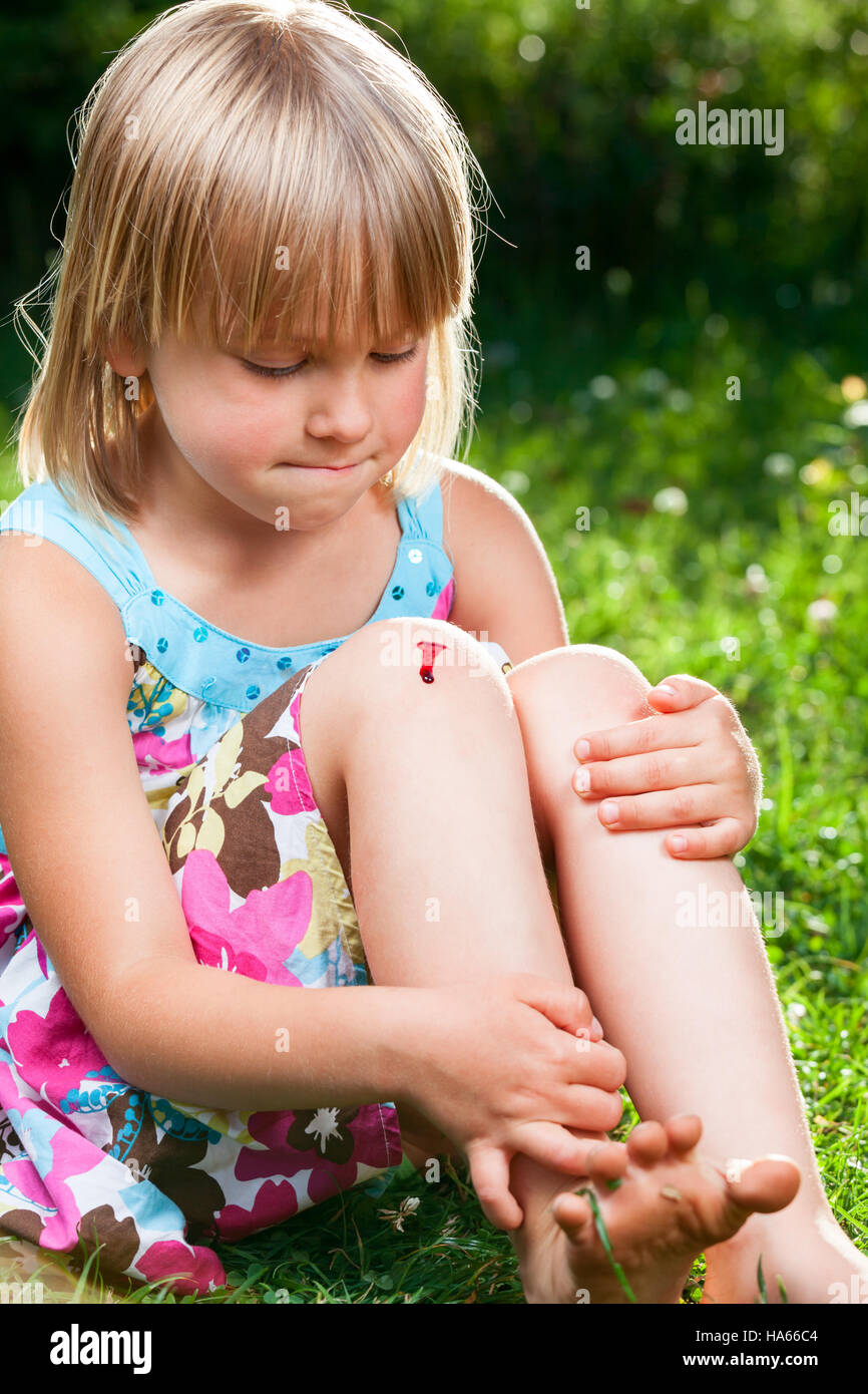 Petite fille assise sur une pelouse dans un jardin d'été à la blessure à son genou avec vos lèvres pincées, retenant les larmes Banque D'Images