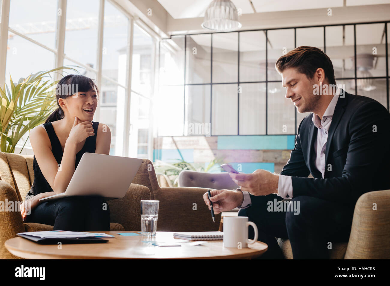 Photo de deux rencontres professionnelles dans le lobby du bureau moderne. Woman working on laptop avec l'homme à l'aide de téléphone mobile. Banque D'Images