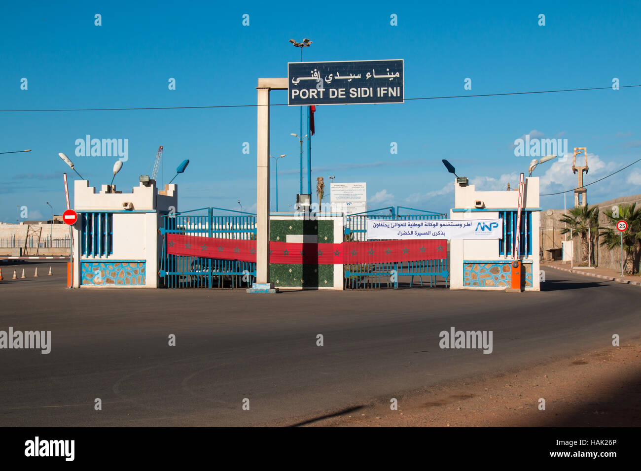 Entrée de la zone d'un port en ville Sidi Ifni à la côte de l'océan Atlantique au Maroc. Clôture avec drapeau marocain. Ciel bleu avec des nuages. Banque D'Images
