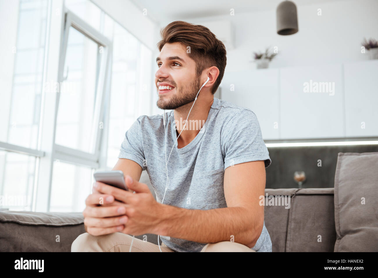 Photo de heureux homme habillé en t-shirt d'écouter de la musique tout en étant assis sur le canapé et de regarder de côté. Banque D'Images