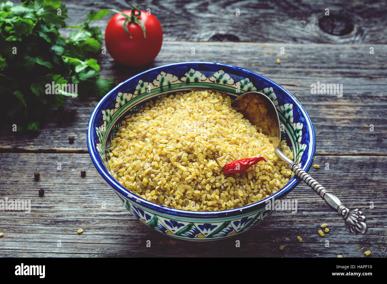 Les grains de blé bulgur brutes dans l'arabe coloré bol, du persil frais, tomates et poivrons pour la cuisson. Arrière-plan de table en bois Banque D'Images