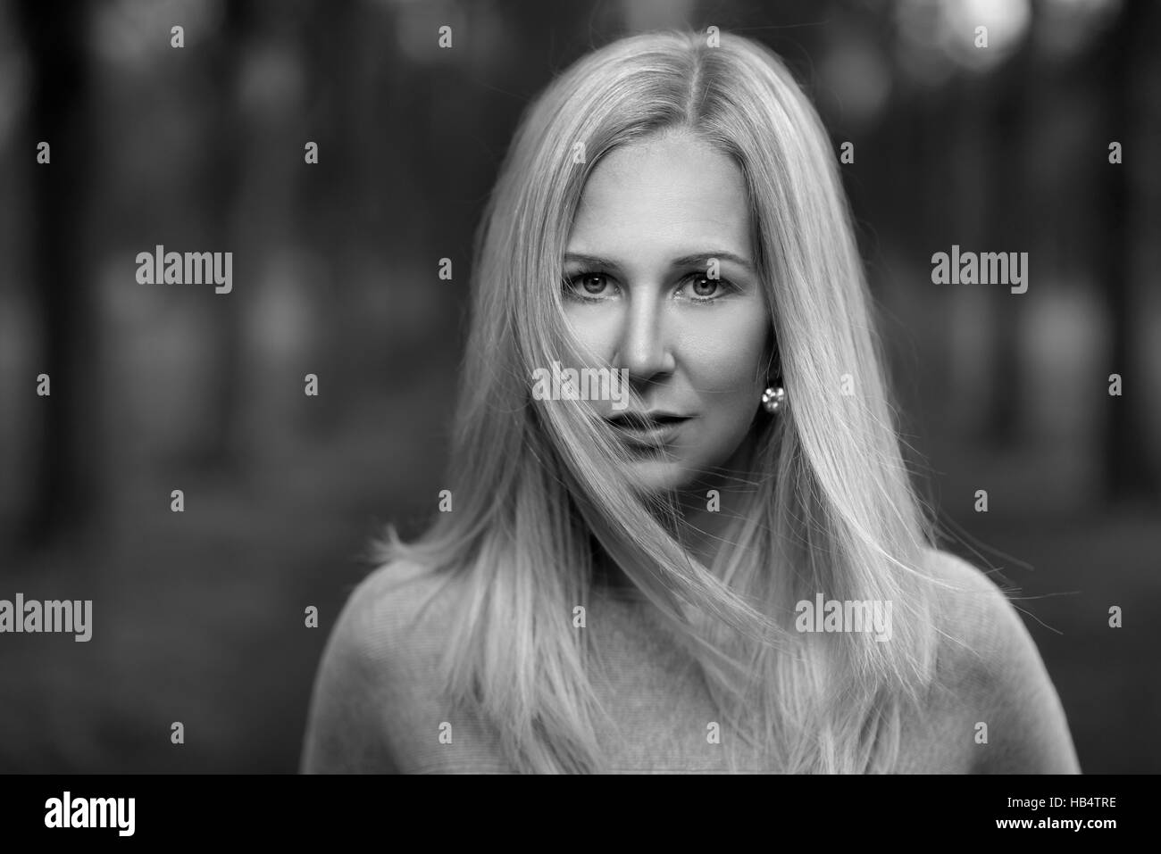 Jolie blonde élégante pensive woman standing outdoors contre une toile de fond des bois avec ses cheveux blonds qui souffle dans la brise à la recherche au camer Banque D'Images