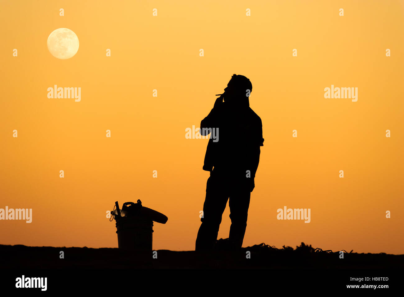 Man smoking cigarette silhouette est un homme debout, fumer une cigarette avec une orange fond coucher de soleil et la Lune se levant dans le ciel. Banque D'Images