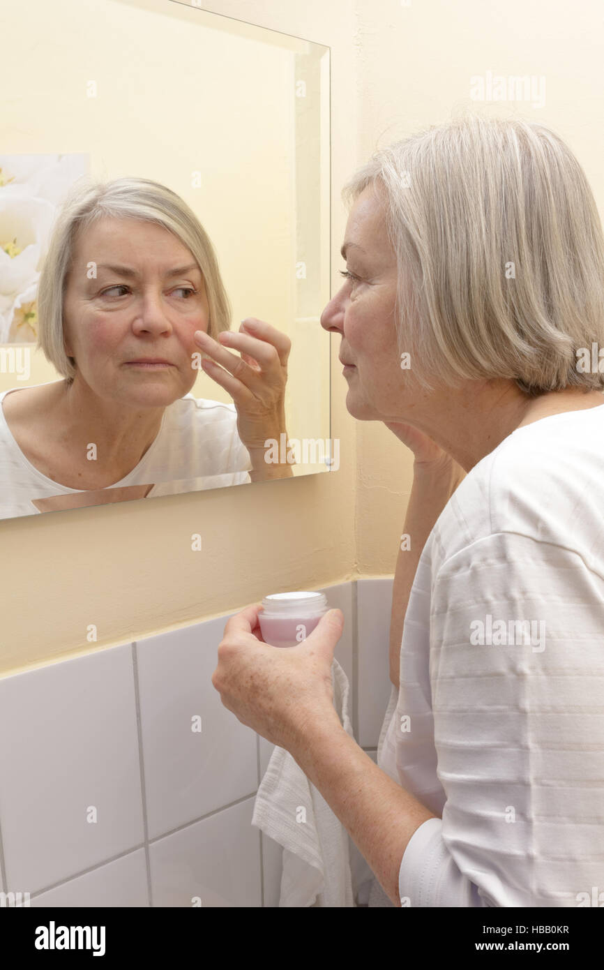 Dame âgée avec une crème pot dans la main, debout devant un miroir dans une salle de bains jaune et l'application de l'hydratant sur son visage Banque D'Images