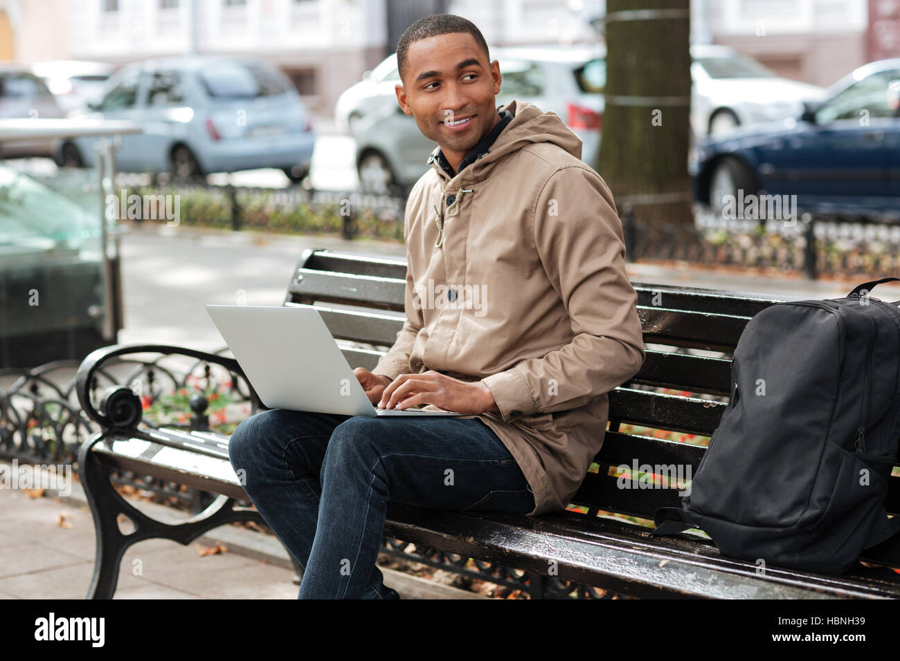 Photo de l'homme heureux avec ordinateur portable assis sur un banc de bois près de sac à dos et la saisie. Regarder de côté. Banque D'Images