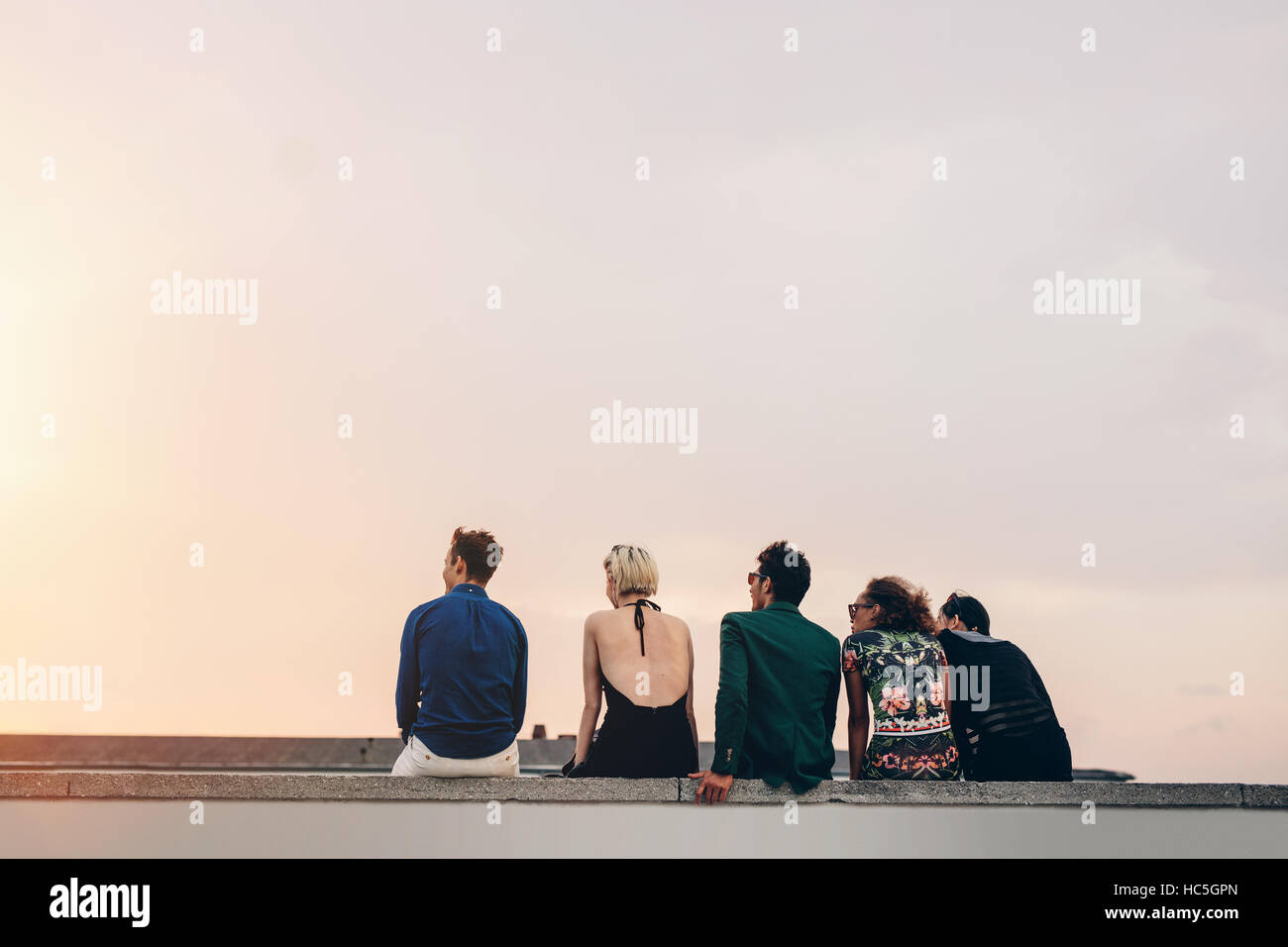 Vue arrière de jeunes amis assis ensemble sur le toit au coucher du soleil. Les jeunes hommes et femmes à traîner sur la terrasse en soirée. Banque D'Images