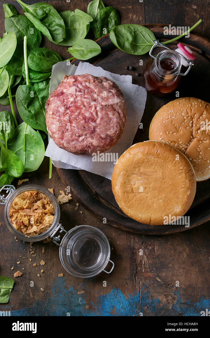 Ingrédients pour faire de hamburger. Escalope de boeuf hamburger cru, deux petits pains, oignon frit, épinards frais et sauce ketchup sur planche en bois sombre plus vieux Banque D'Images