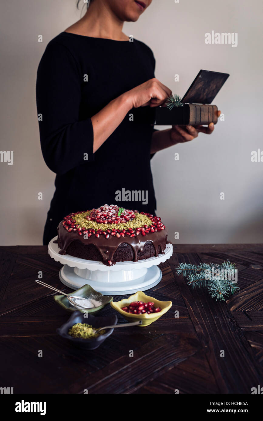 Une femme tient une boîte cadeau derrière une table où un gâteau au chocolat de Noël et trois petits bols avec graines de grenade, de la poudre de pistache et sug Banque D'Images