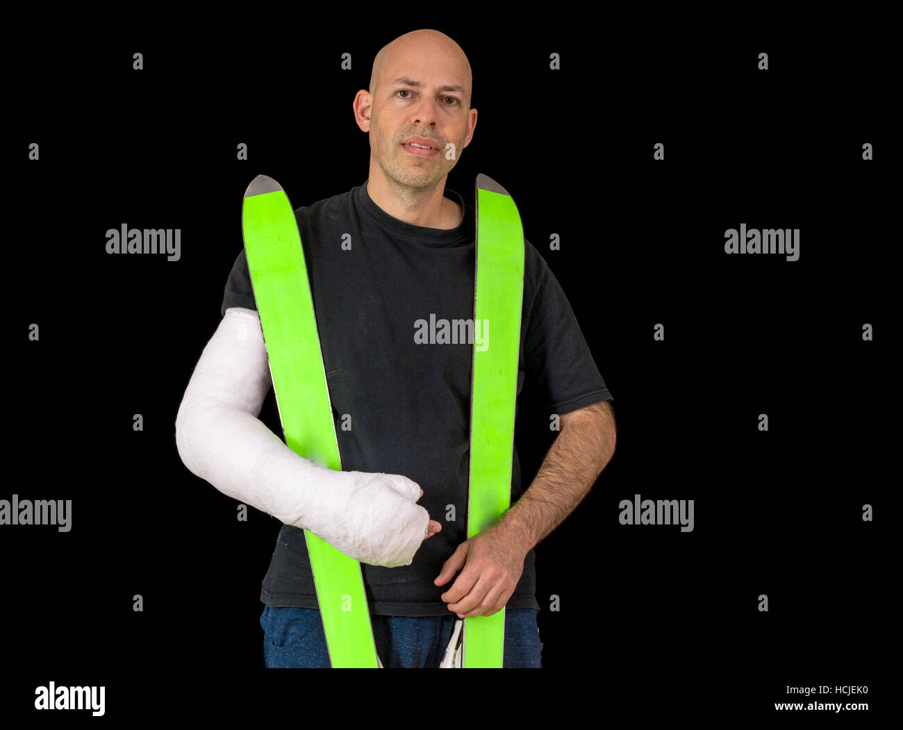 Jeune homme portant un bras blanc brillant, en tenant ses skis après avoir cassé le poignet dans un accident de ski alpin, isolé sur fond noir Banque D'Images
