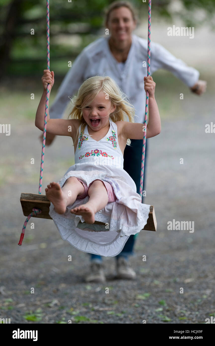 Blonde, pieds nus petite fille portant une robe d'été est poussée par sa mère sur une balançoire sur une journée d'été. Banque D'Images