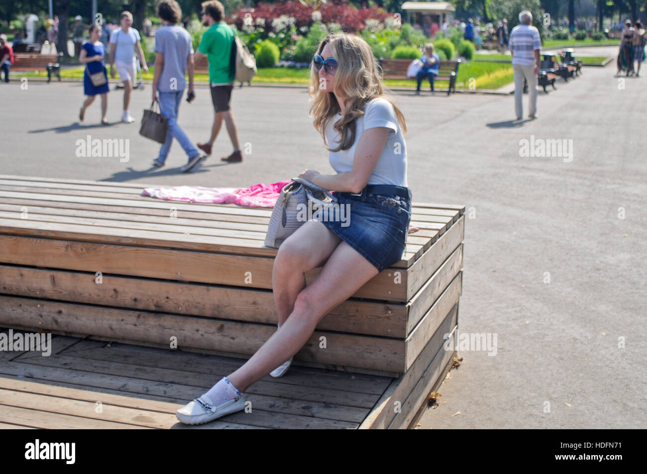 Femme blonde portant des lunettes de soleil et un Jean mini jupe, assise sur un banc en bois. Parc Gorky, Moscou Banque D'Images