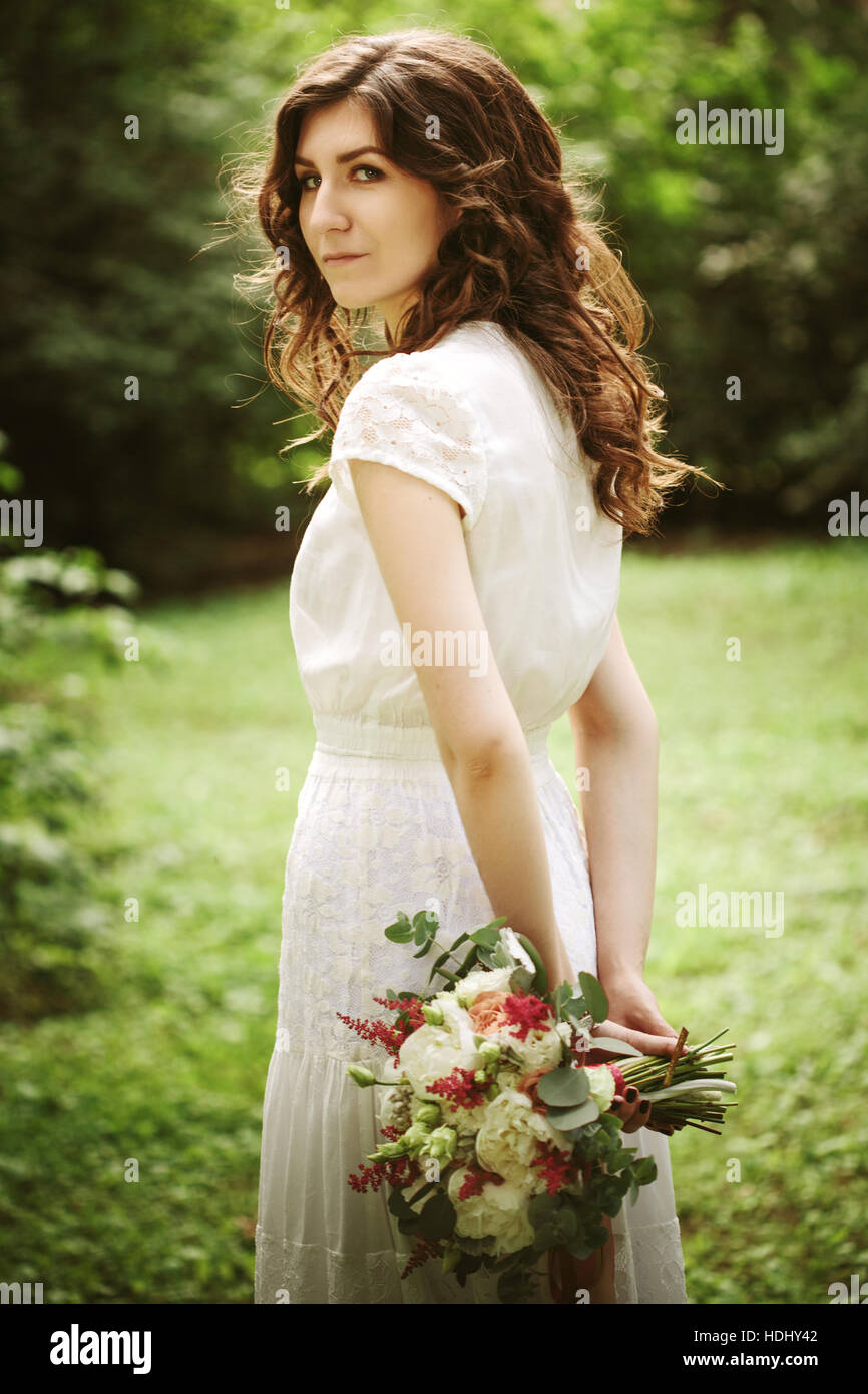 Jeune mariée dans le parc tenant des fleurs de mariage derrière son dos, looking at camera Banque D'Images