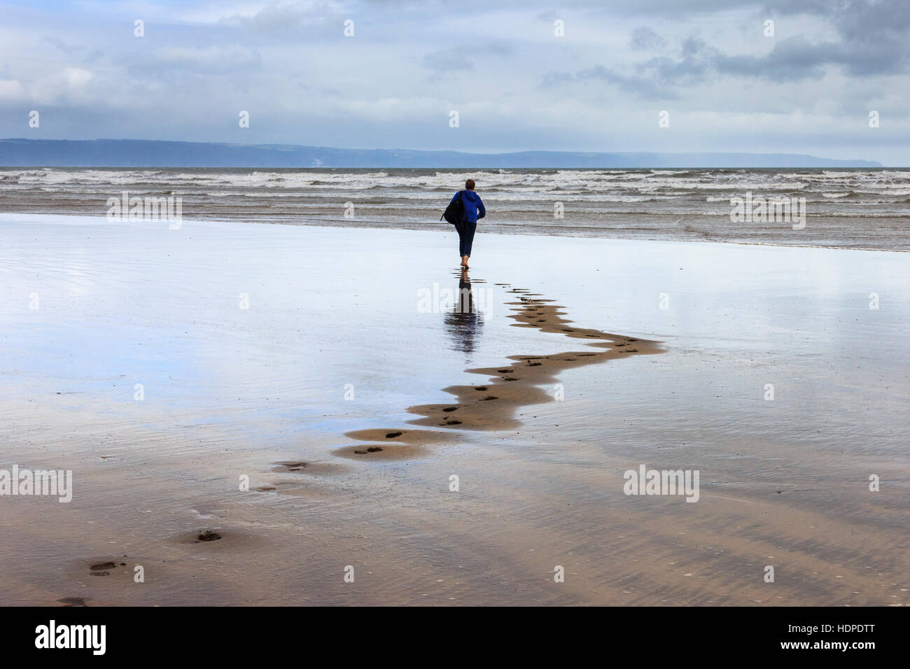 Une femme marchant pieds nus vers la mer sur une plage balayée par le vent laissant des traces de pas dans le sable, Westward Ho !, Devon, England, UK Banque D'Images