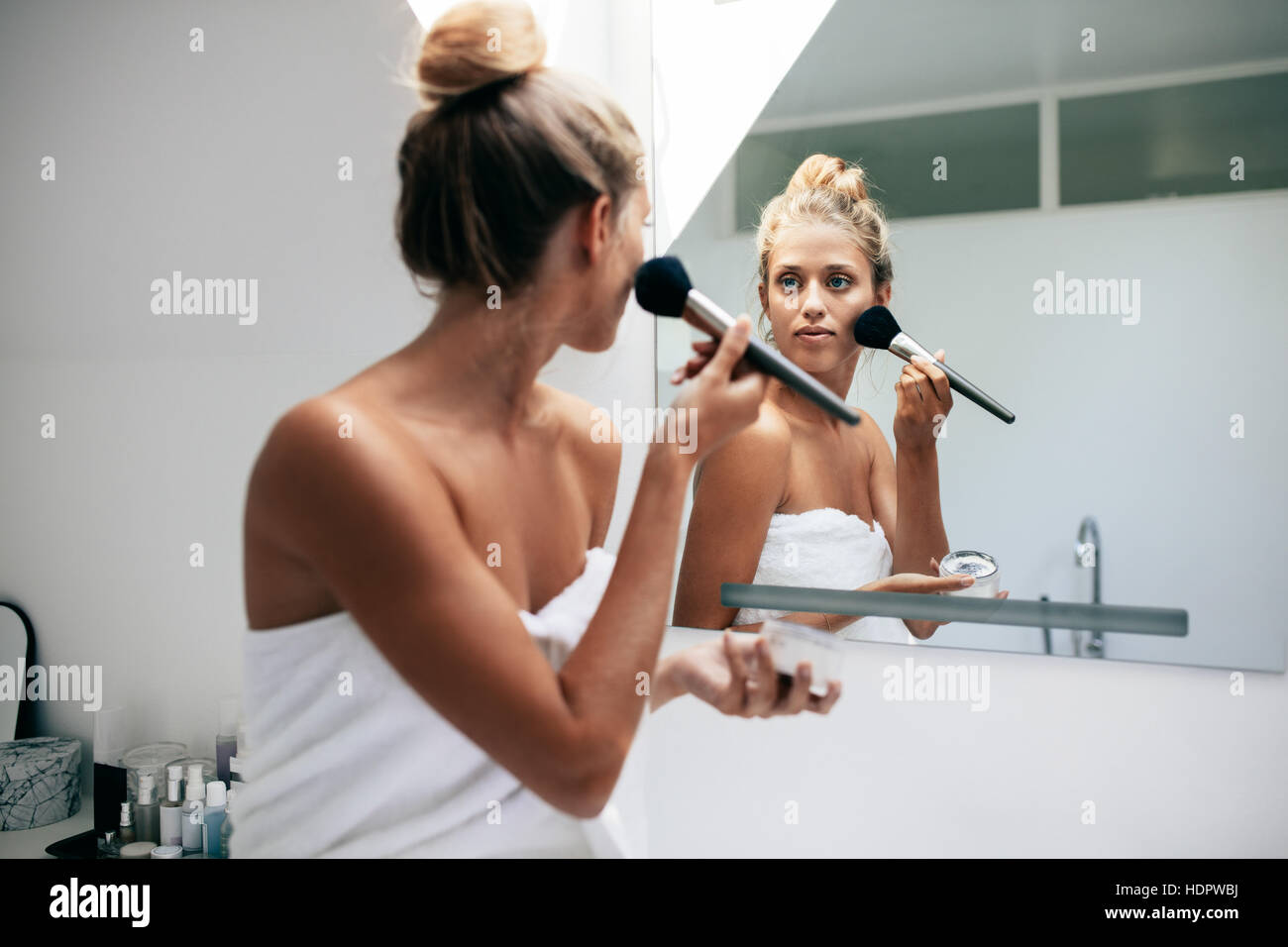 Belle femme d'appliquer des cosmétiques sur son visage dans la salle de bains. Femme à la recherche dans le miroir de salle de bains et la mise sur le maquillage. Banque D'Images