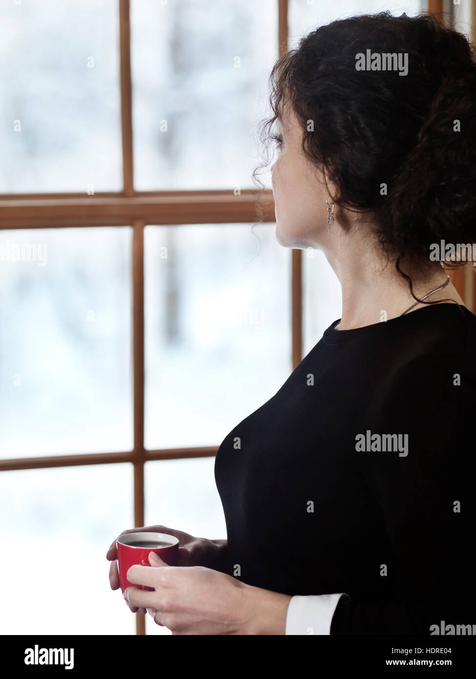 Femme avec une tasse de café en regardant par la fenêtre avec un paysage d'hiver enneigé derrière elle Banque D'Images