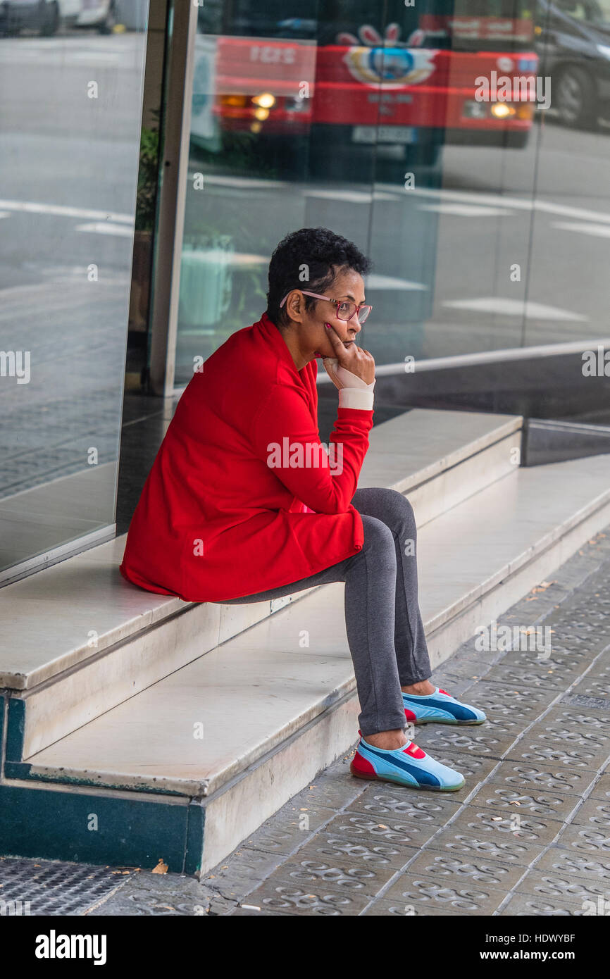 Une femme espagnole vêtu d'une veste rouge et des chaussures de course de  fantaisie et collants, est assis sur des marches en marbre à l'extérieur  d'attente avant un magasin à Barcelone, Espagne
