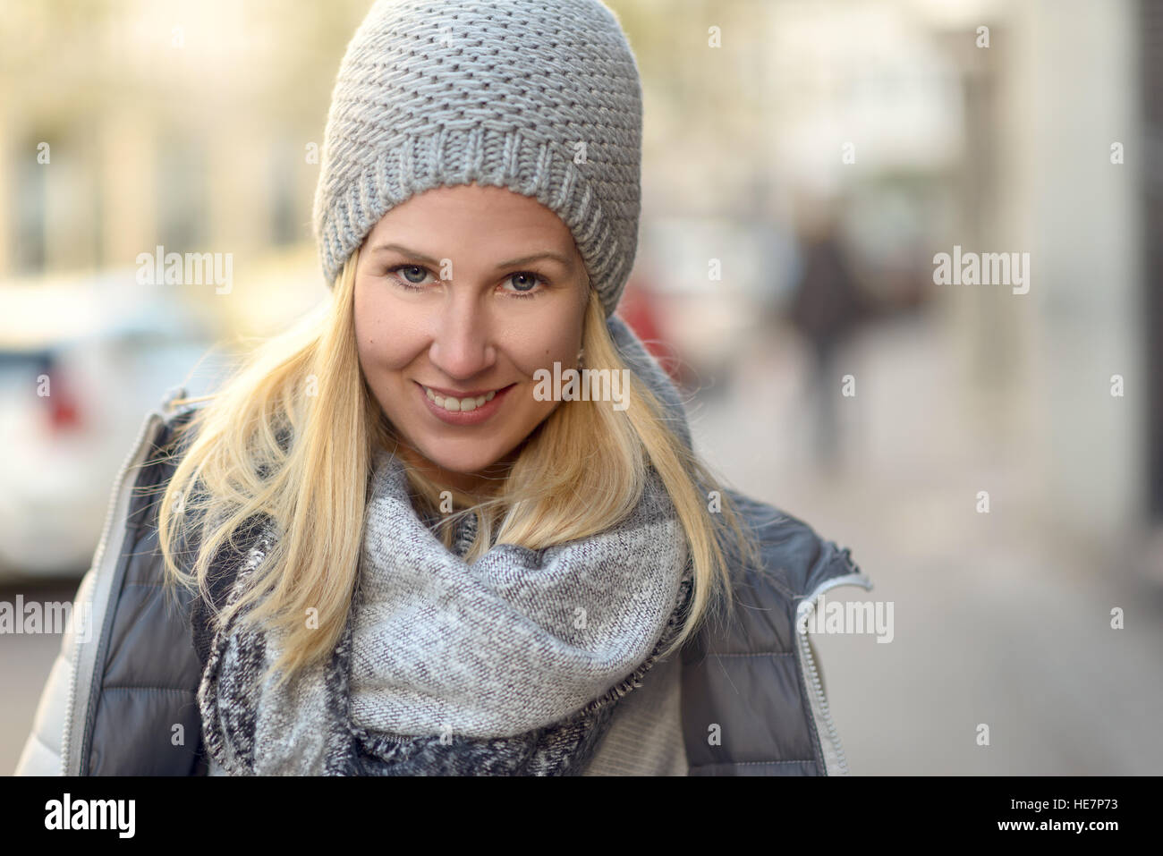 Friendly Smiling attractive young woman in un hiver gris élégant costume avec Chapeau tricoté et écharpe et veste chaude dans une rue urbaine Banque D'Images
