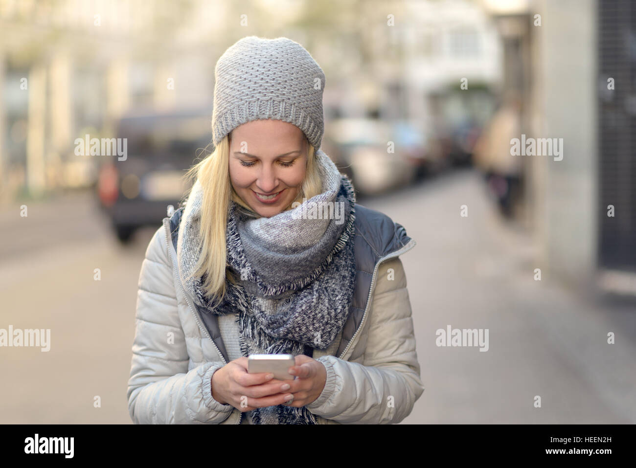 Jeune femme à la mode en gris Down jacket et écharpe occupé avec son téléphone mobile en marchant une rue de la ville Banque D'Images