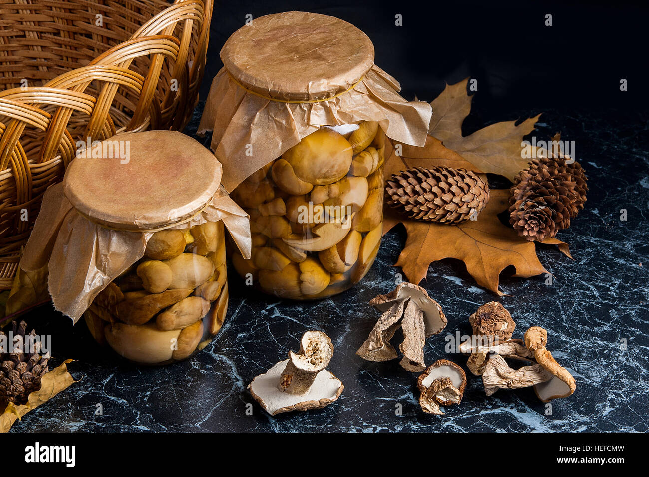 Deux pots en verre avec des champignons marinés sur noir en arrière-plan. Plusieurs champignons blanc sec sur la table. Panier en bois jaune à l'arrière Banque D'Images