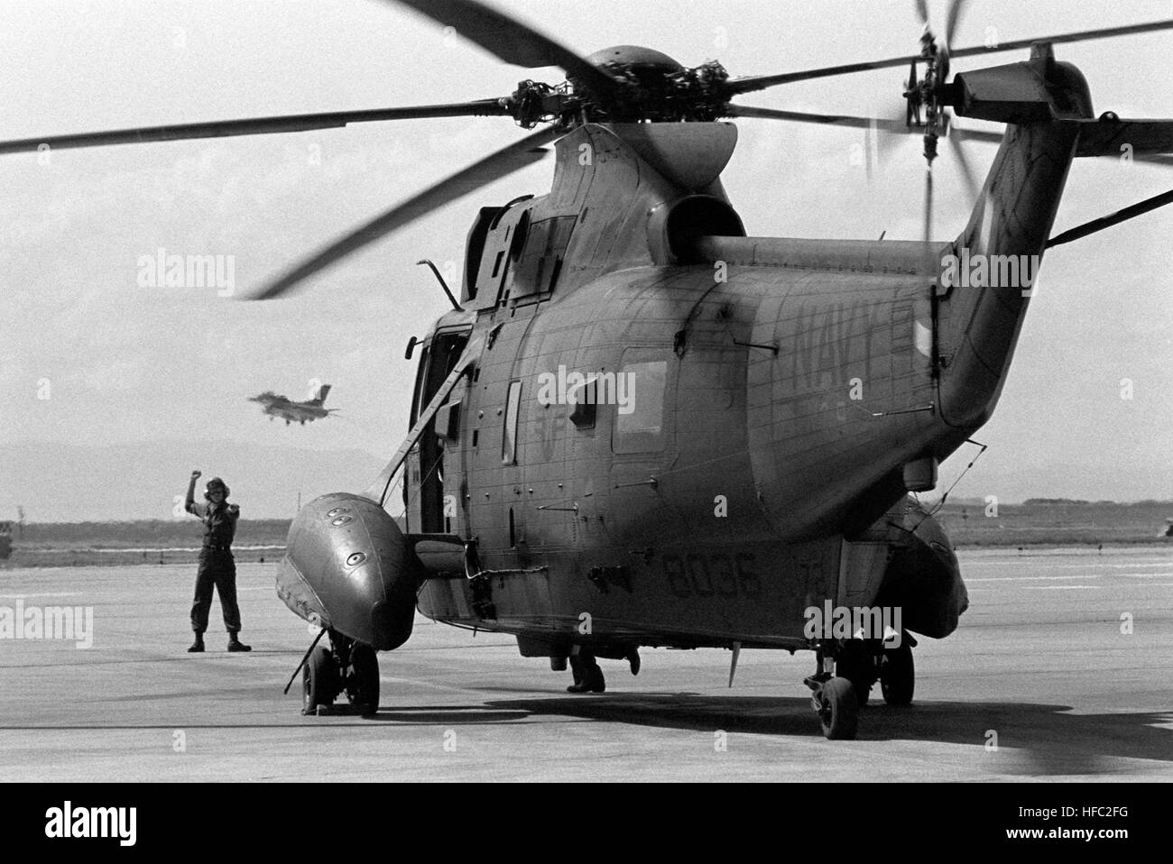 La Marine américaine (USN) Sikorsky SH-3 Sea King se trouve sur l'axe de vol comme un motif d'équipage signale au pilote pour décoller lors d'une recherche et sauvetage (SAR) à l'exercice Naval Air Station (NAS) l'île de Whidbey, Washington (WA). L'US Air Force (USAF) General Dynamics F-16 Fighting Falcon est vu sur approche à l'atterrissage dans l'arrière-plan. HH-3A SC-9 à l'île de Whidbey NAS 1983 Banque D'Images