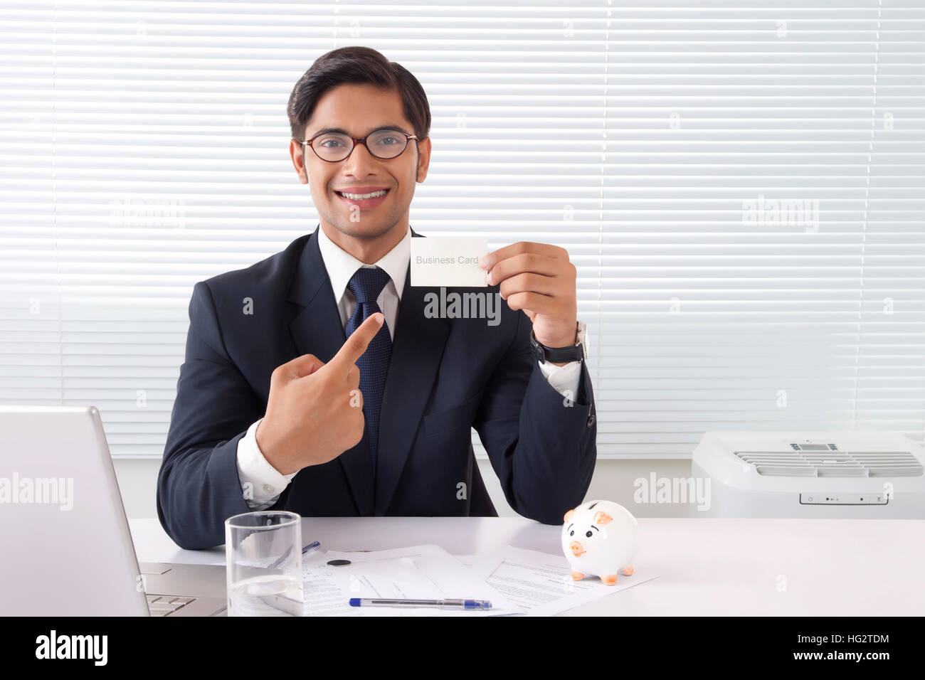 Jeune homme professionnel montrant carte d'affaires au bureau avec ordinateur portable et piggy bank on table Banque D'Images