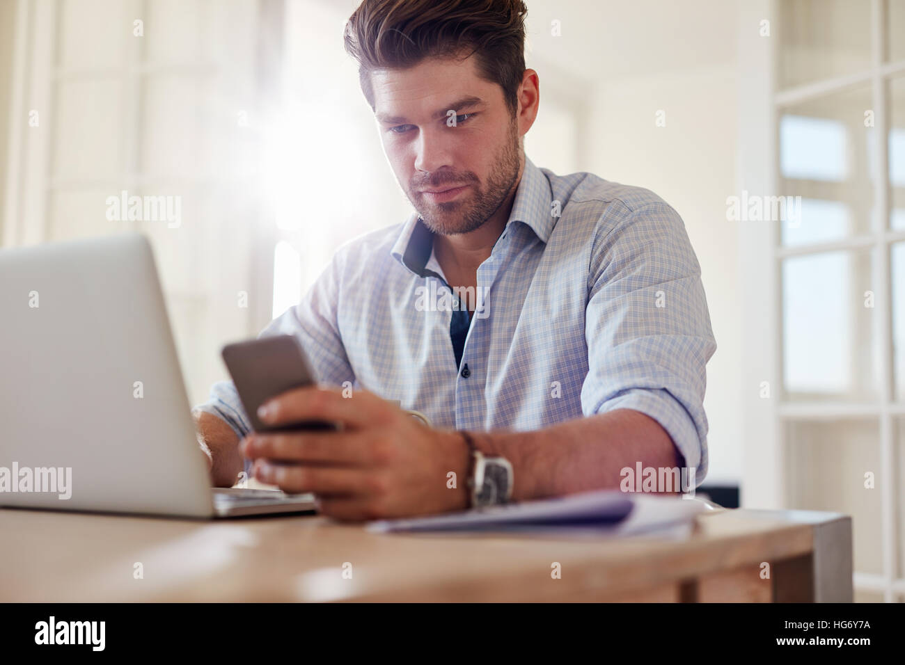 Shot de confiance man reading text message sur son téléphone portable tout en travaillant sur l'ordinateur portable. Jeune homme travaillant à la maison. Banque D'Images