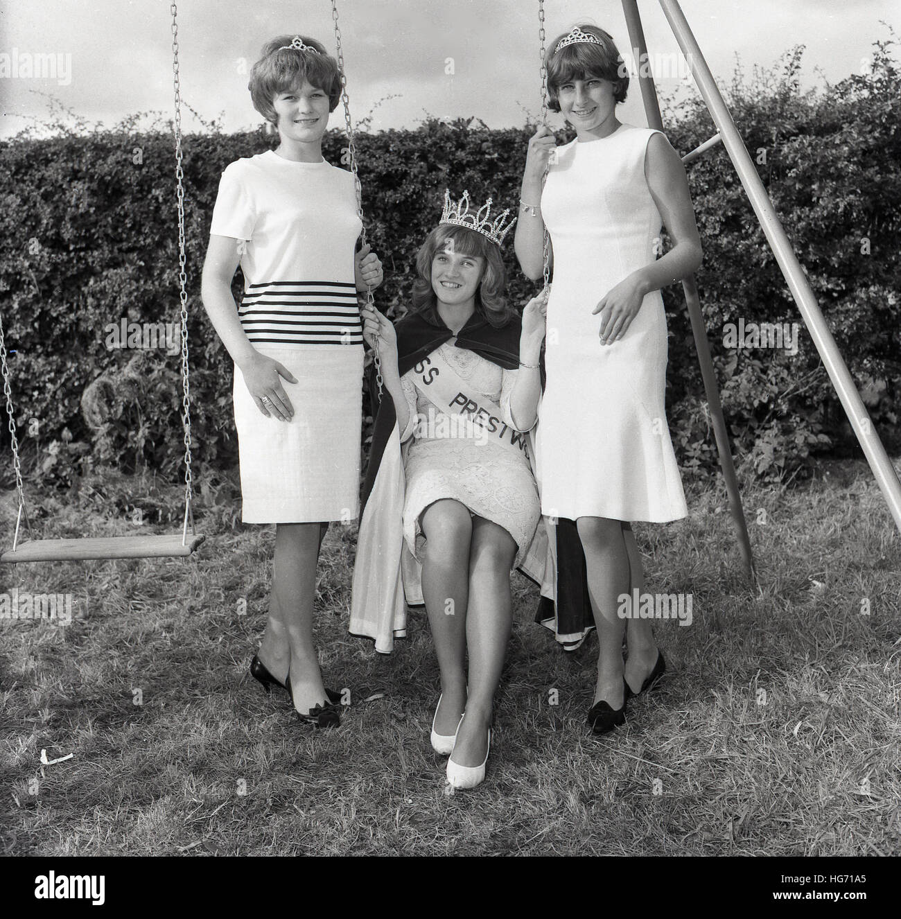 Années 1960, historiques, vainqueur de l'entre 2 lacs 'Miss' concours de beauté assis sur une balançoire avec sa couronne, avec ses deux finalistes debout à côté d'elle, England, UK. Banque D'Images
