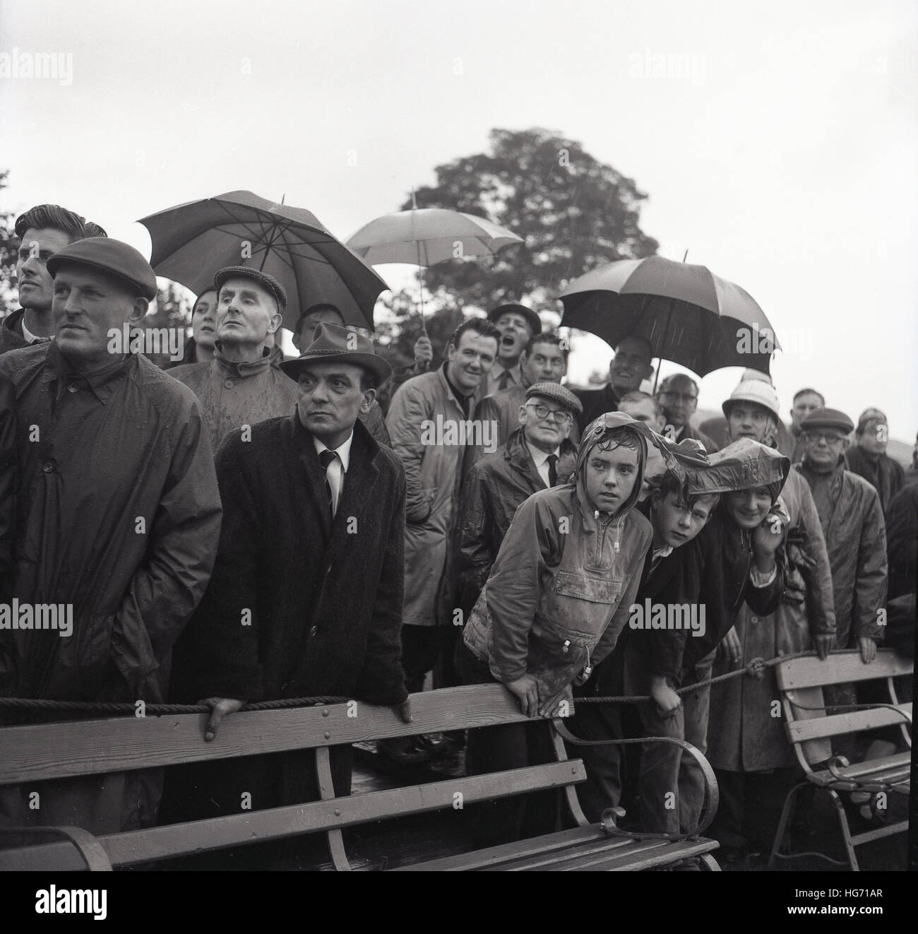 1965, historique, un groupe de spectateurs masculins dans les imperméables et certains portant des casquettes et des parasols, debout regardant un match de foot sous la pluie, England, UK. Banque D'Images