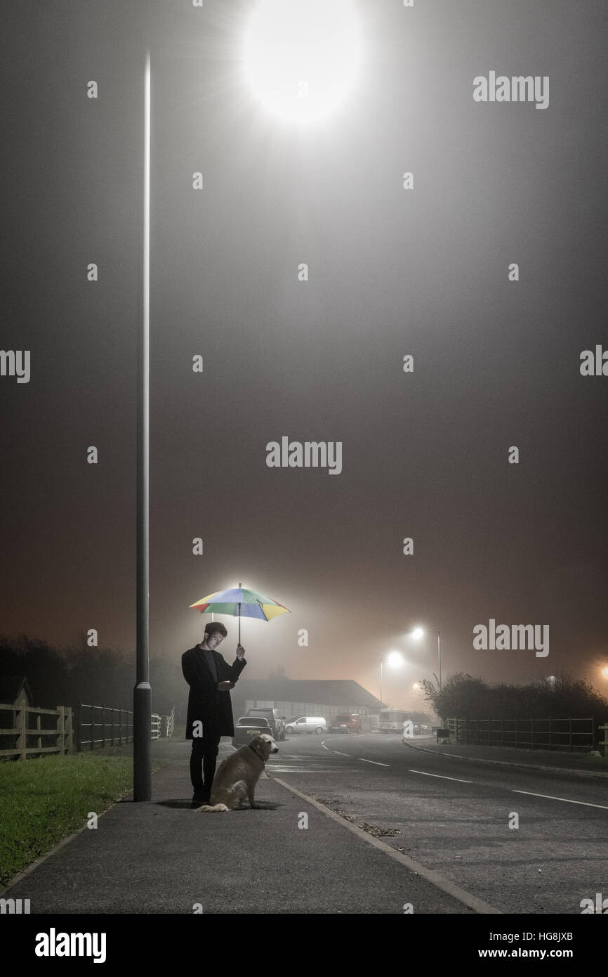 Adolescent sur téléphone mobile avec chien sous l'éclairage dans le brouillard avec parapluie Banque D'Images