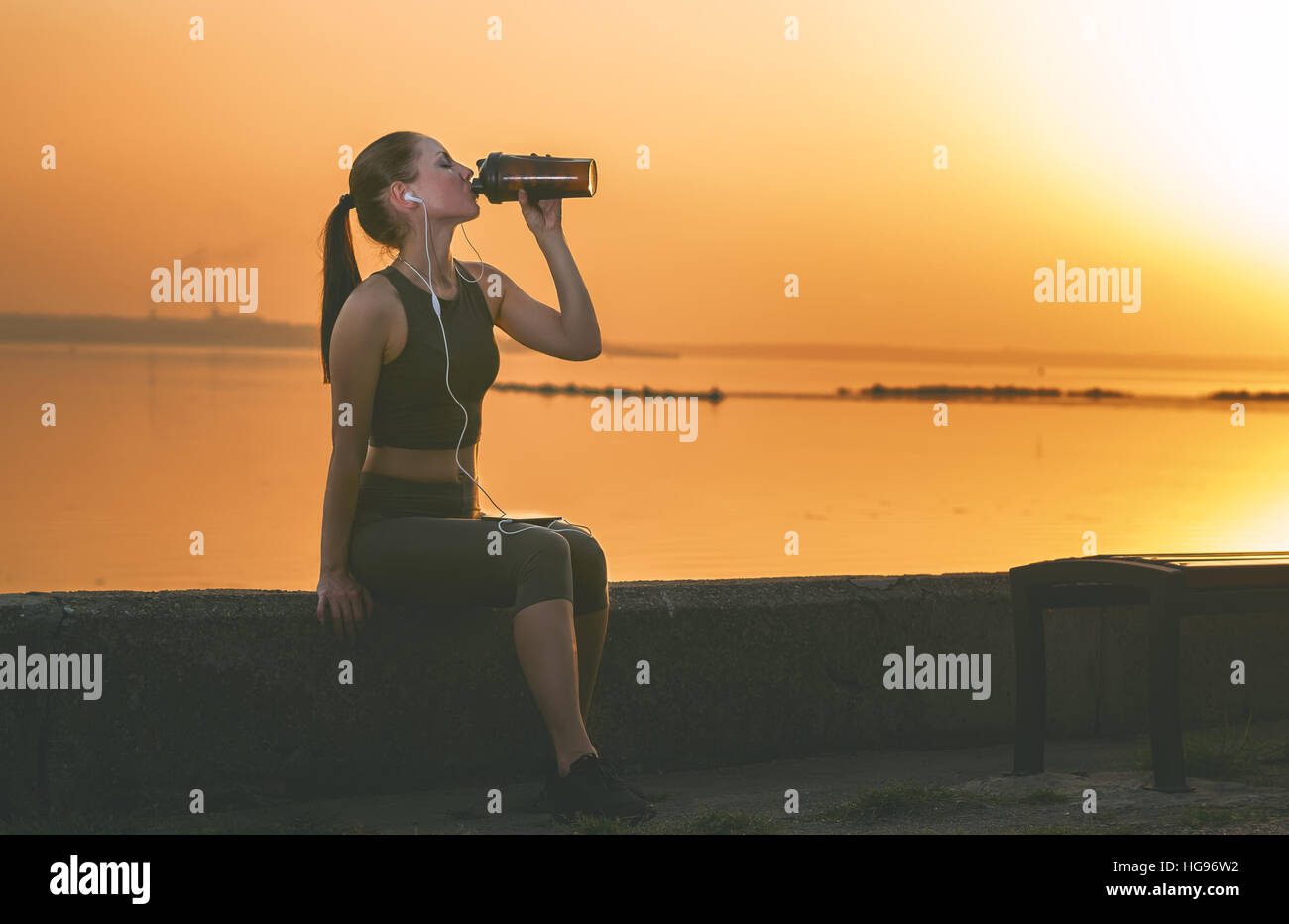 Jeune fille après le jogging. La sportive de boissons un shaker dans le parc sur un fond de coucher du soleil orange. Banque D'Images